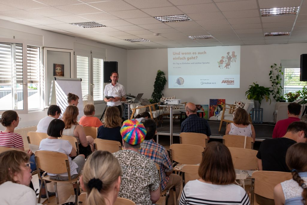 Foto vom Tag, auf dem der Raum mit den Teilnehmenden, dem Moderator und der Präsentation zu sehen ist